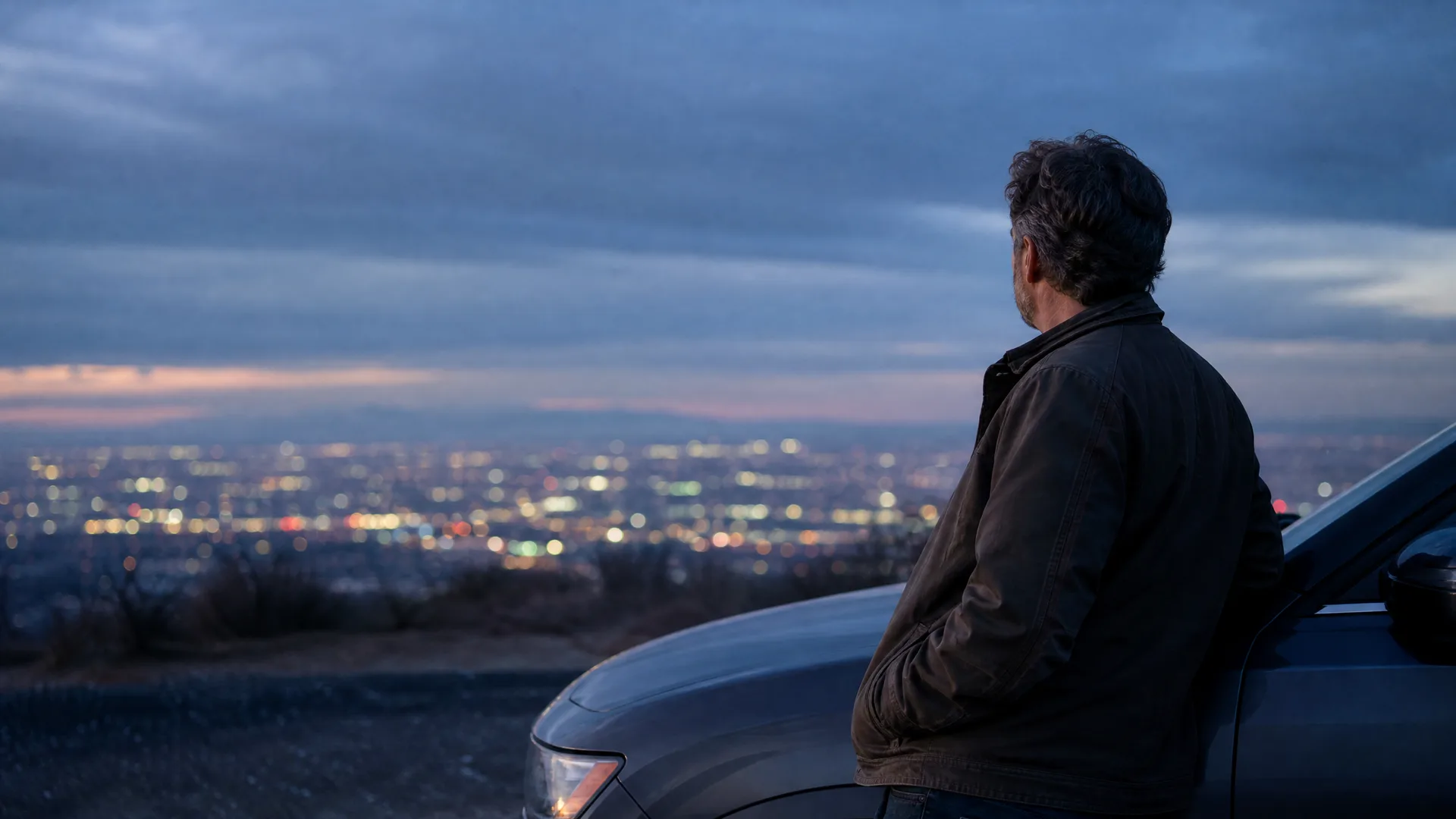 A man in a dark jacket leans against the front of a vehicle on a hillside at dusk, looking out over a blurred city skyline of lights.