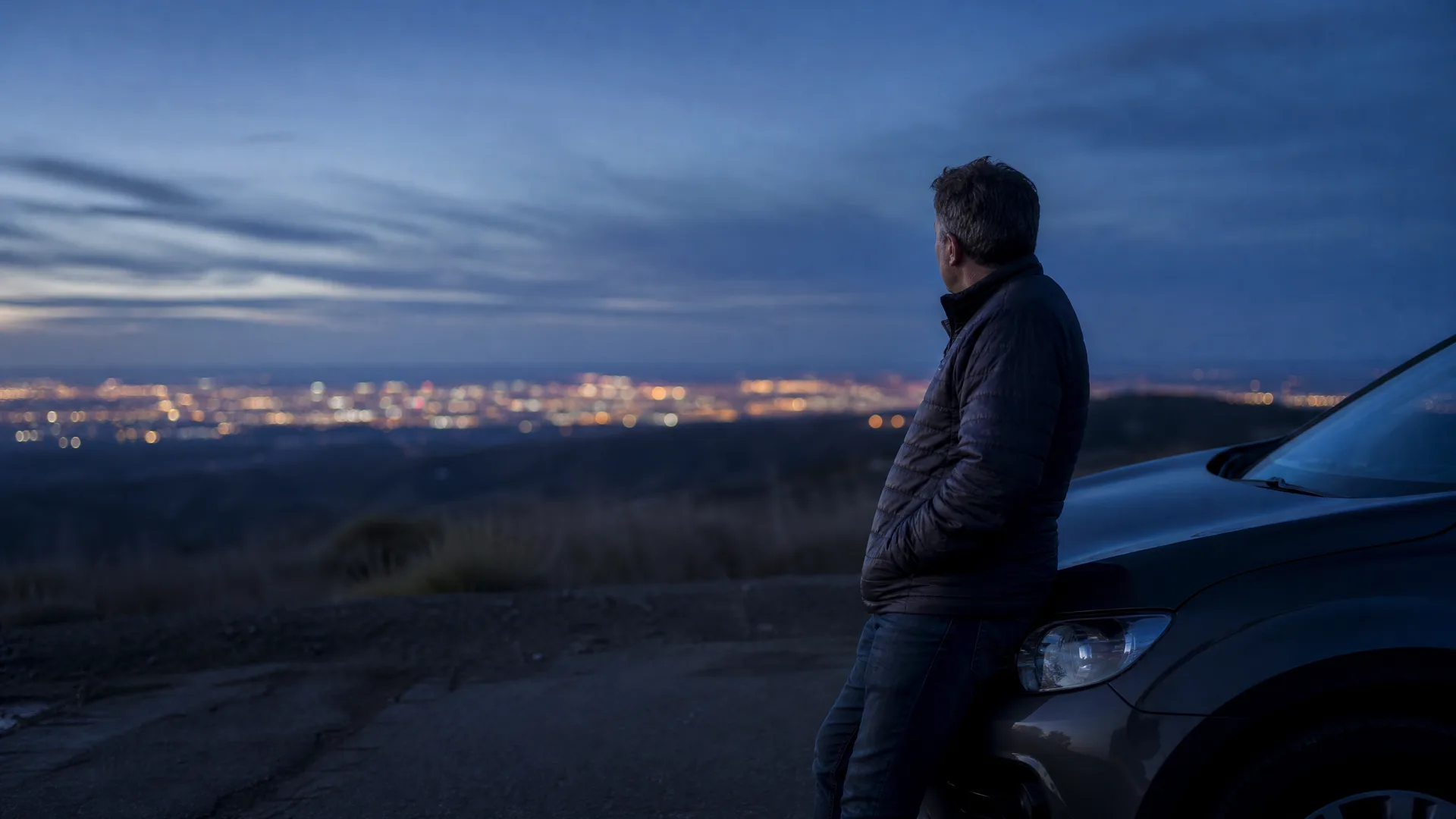 A man in a dark quilted jacket leans against the front of a car on a dirt pullout at dusk, looking out over distant city lights below.