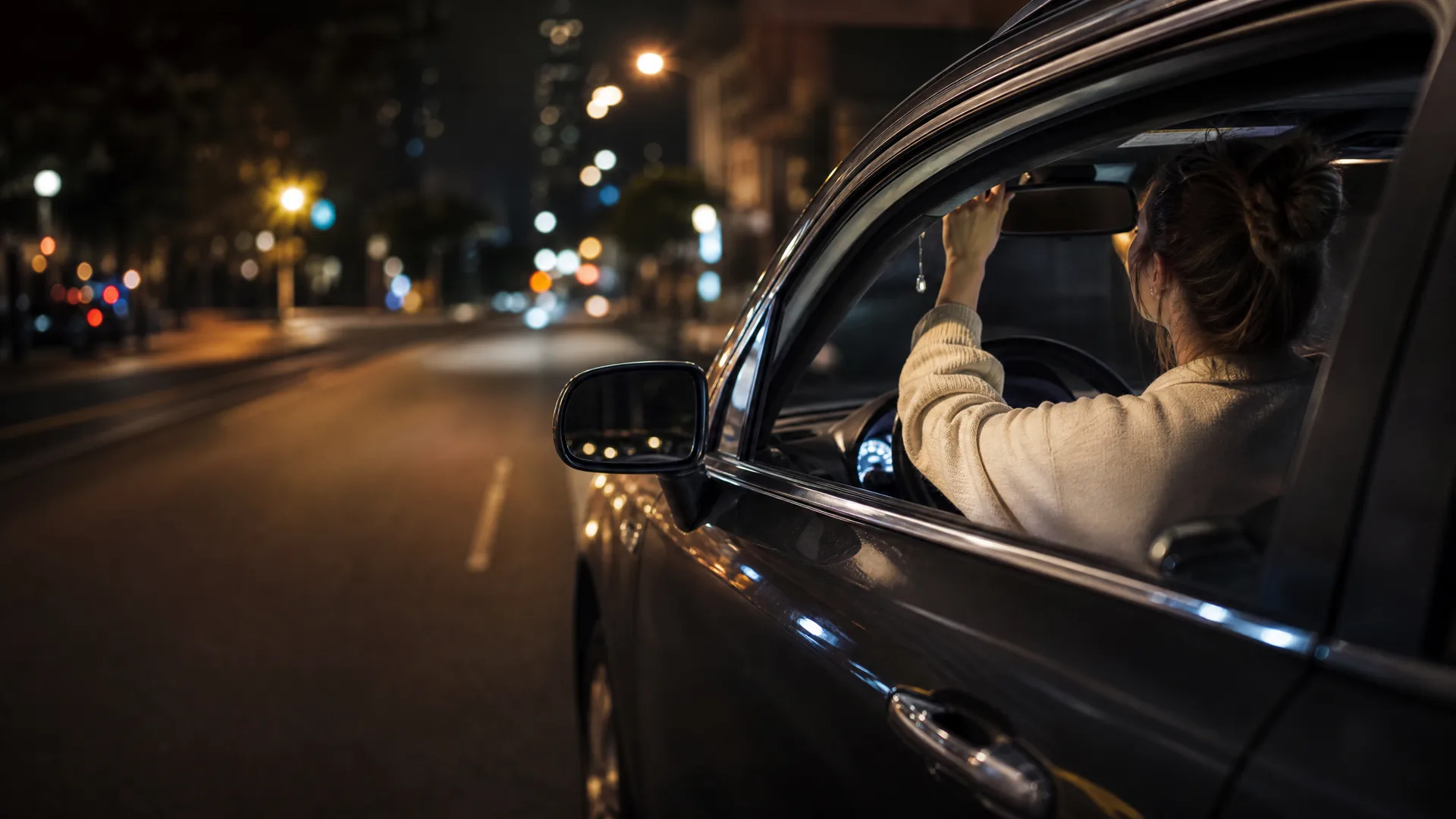 A woman in a light sweater sits in the driver's seat of a car at night, seen from outside through the open window, with blurred city street lights ahead.