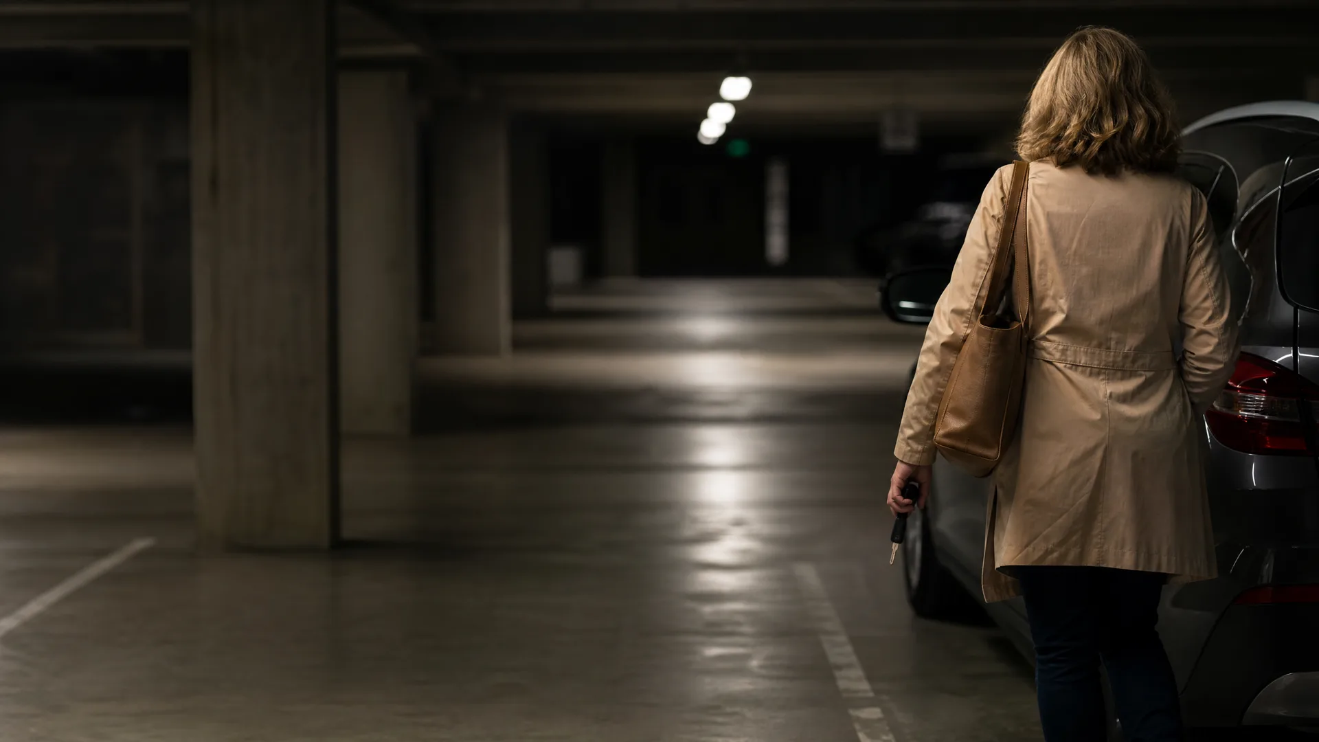 A woman in a beige coat with a shoulder bag walks away from the camera toward a parked car in a dimly lit underground parking garage with concrete columns.