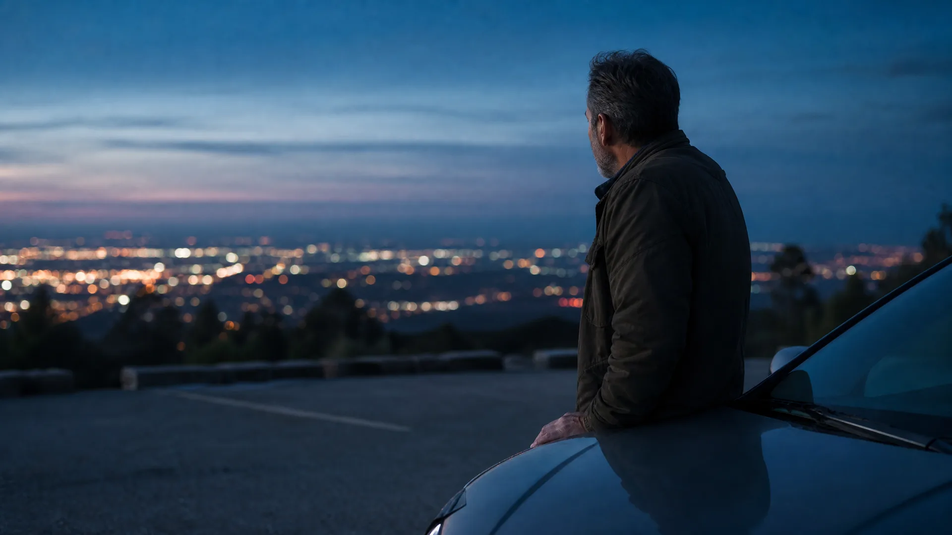 A man in a dark jacket leans against the hood of a car in a hilltop parking area at dusk, looking out over distant city lights.