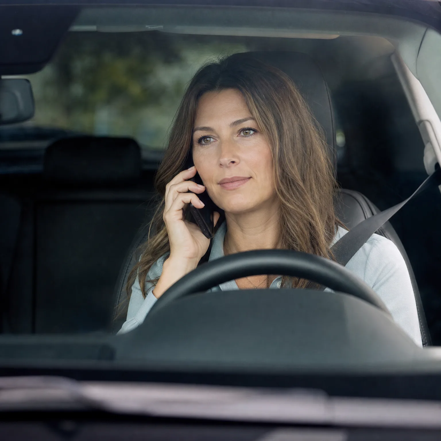 A woman with long brown hair sits in a car's driver seat holding a phone to her ear, seen through the windshield above the steering wheel.