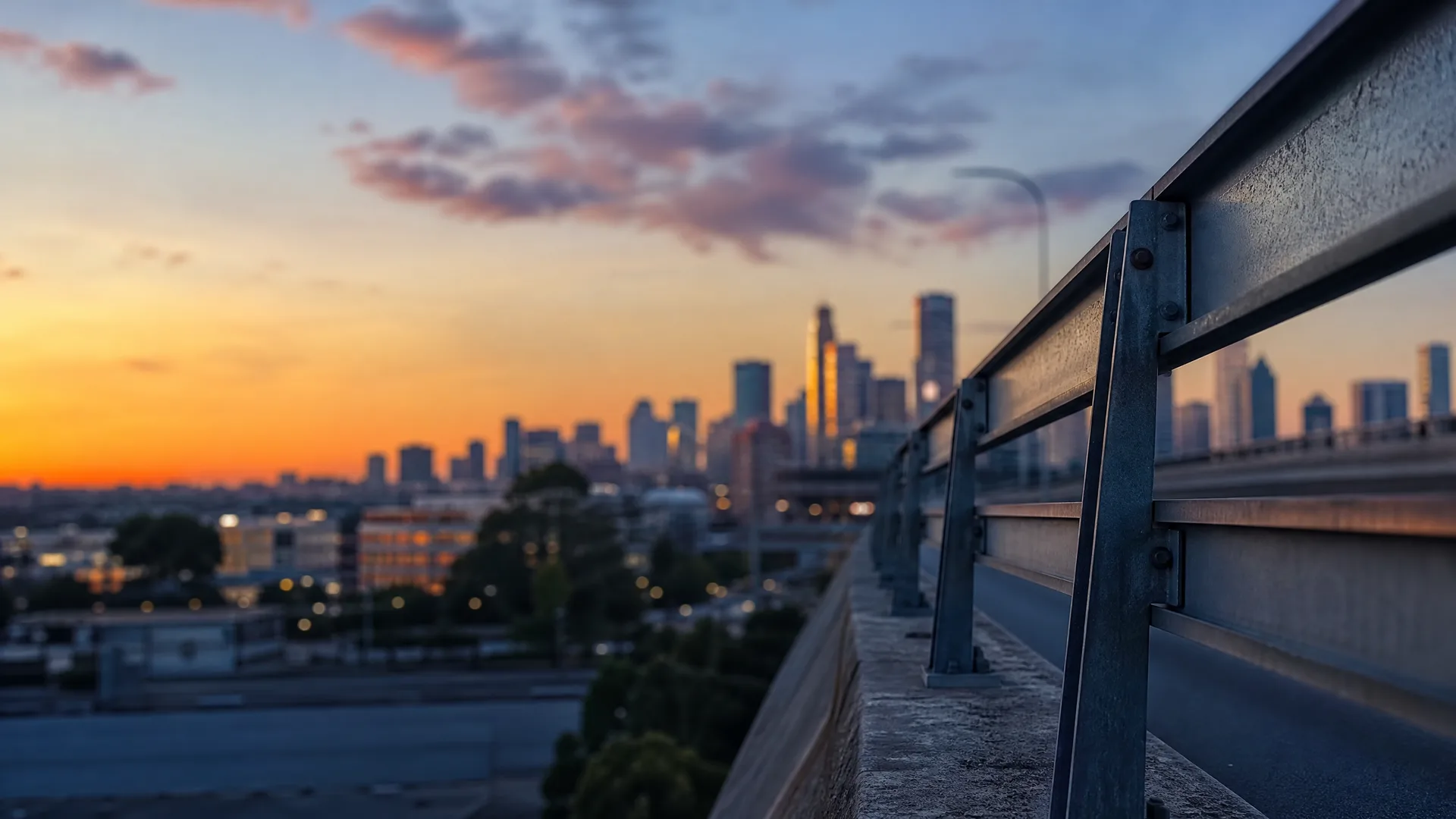 A metal guardrail along an elevated roadway in sharp foreground focus, with a blurred city skyline and orange sunset sky behind.