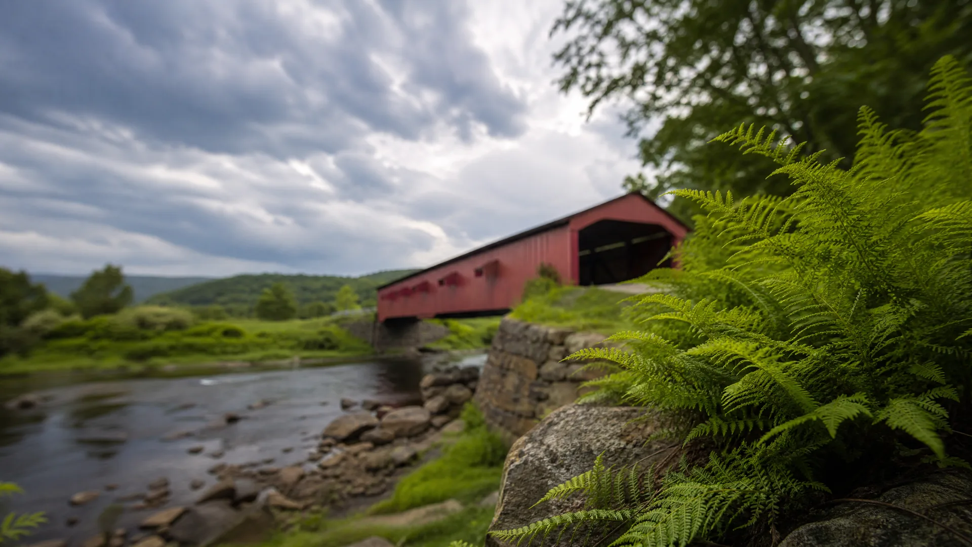 A red covered wooden bridge on stone abutments spans a shallow rocky river, with ferns in sharp focus in the foreground and cloudy sky above low green hills.